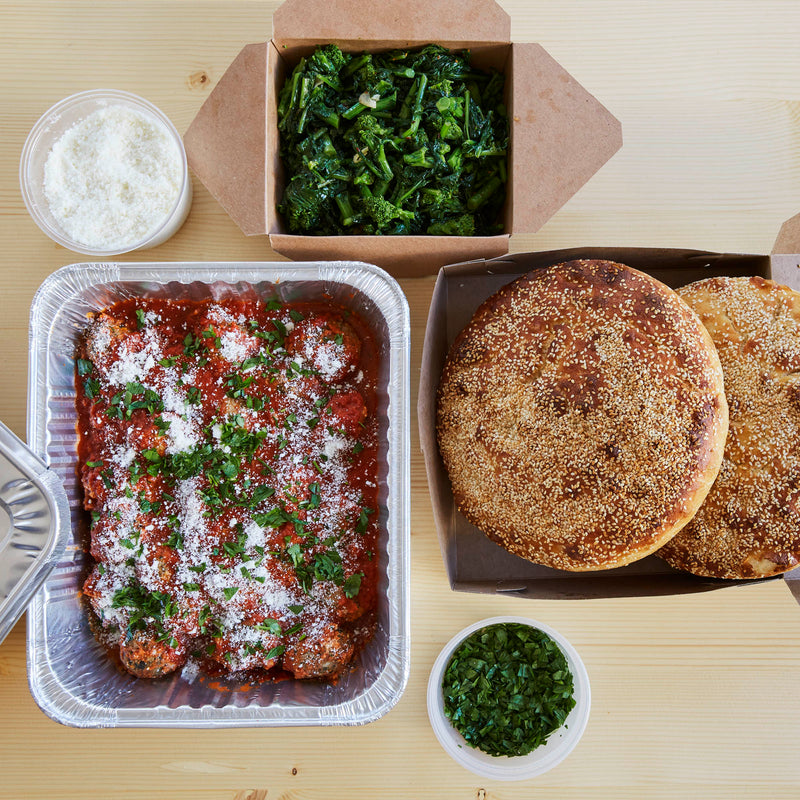 A takeout meal featuring meatballs in sauce with parmesan, sautéed greens, large bread rolls, and grated cheese.