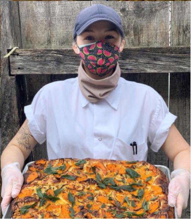 A masked woman holds out a pizza.