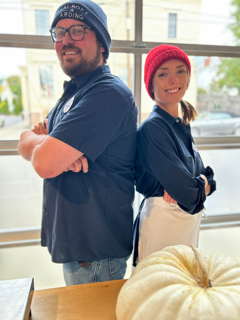 Two smiling people standing back to back, wearing beanies.