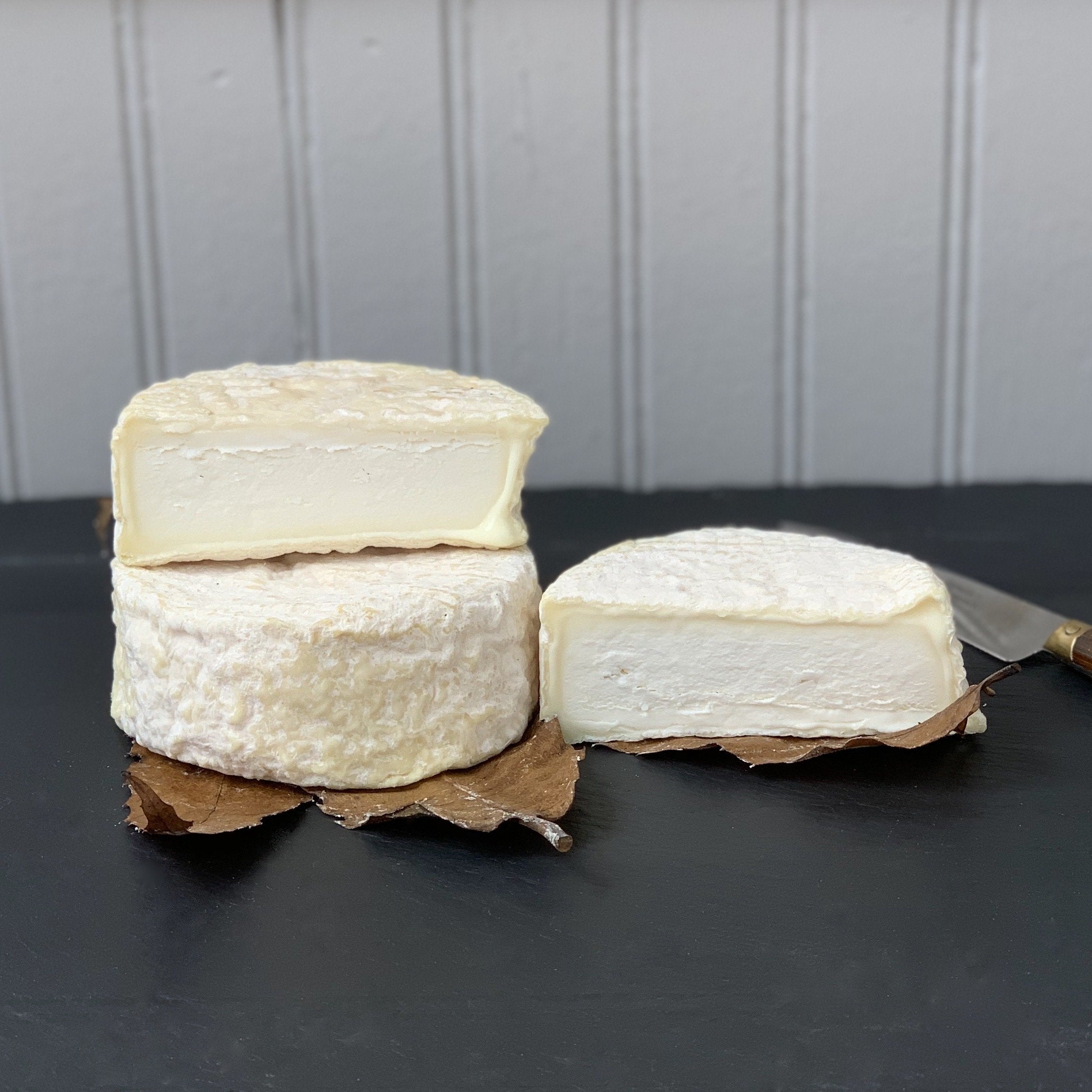 Two wheels of soft cheese on a black surface with one cut in half.